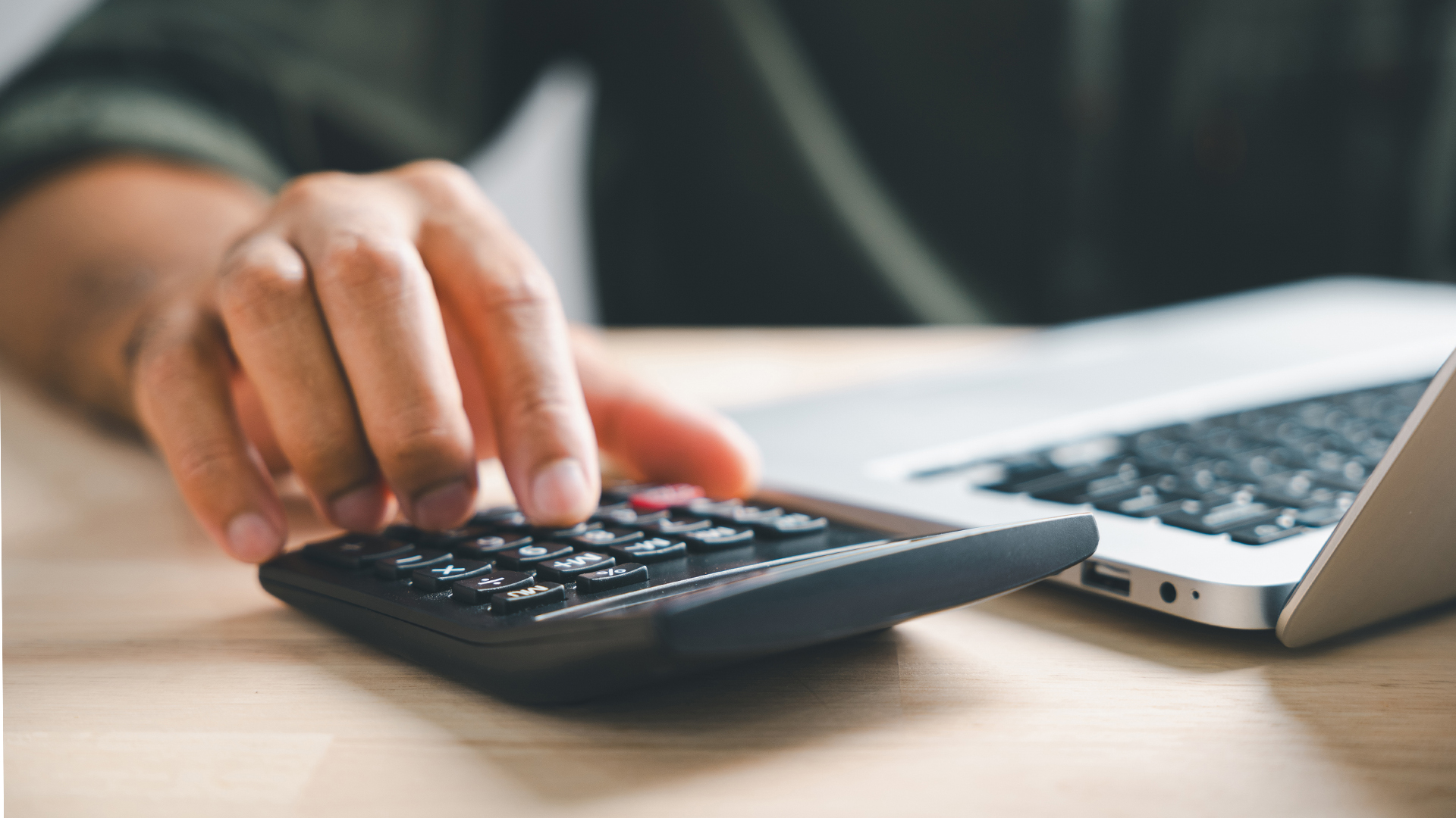 Hand on calculators, aiming for home refinance. Wooden house model, buy or rent note on desk.
